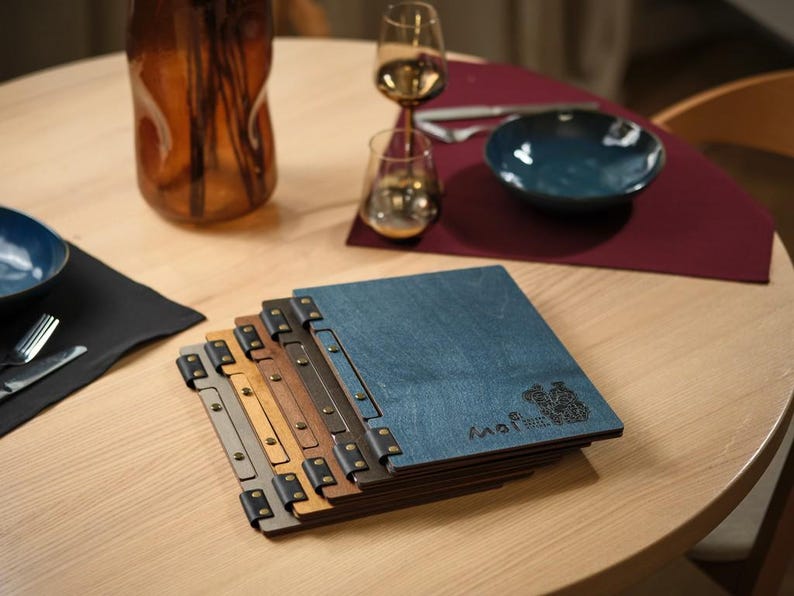 Grey stone-textured square menu cover on a warm wooden table with an amber glass vase and a dark ceramic bowl in the background. Metal hinge hardware on the spine. Closed. The warm props contrast pleasantly with the cool grey cover surface. The amber glass and dark ceramics feel Japanese in their simplicity, which reinforces the overall aesthetic. The cover looks at home in this setting — understated, considered, nothing competing with it for attention on the table surface.