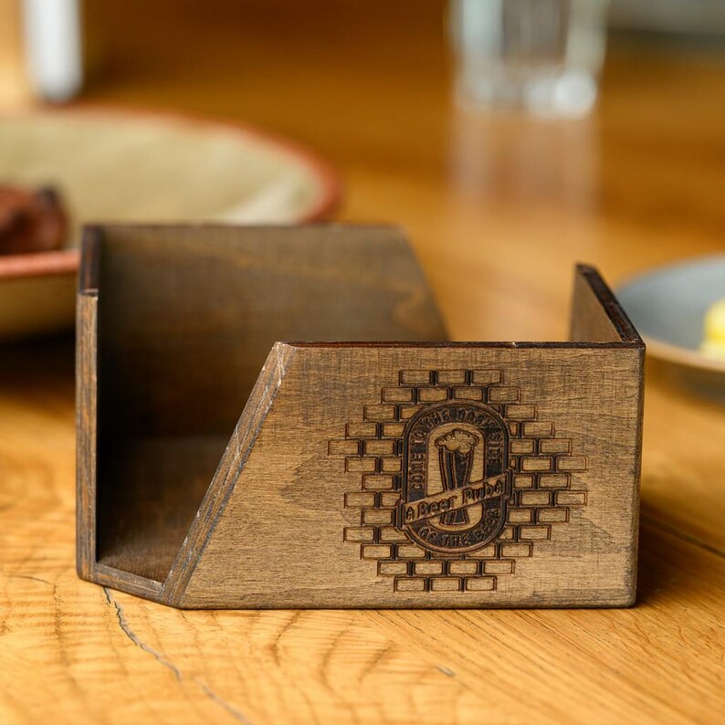 Close-up of a dark walnut-stained plywood angled napkin holder with engraved pub logo holding white paper napkins on a rustic wooden bar table. The open-top access design and dark finish are shown close in an authentic bar dining setting. Custom engraved wooden napkin holder for pub table service, craft beer bar dining rooms, restaurant interiors, and hospitality branding with personalized plywood woodcraft table accessories.