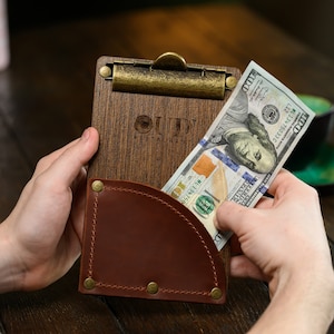 May include: A wooden clipboard with a brass clip and a brown leather pocket. The clipboard has the word "OUD RESTAURANT" printed on it. A hand is holding a one hundred dollar bill.