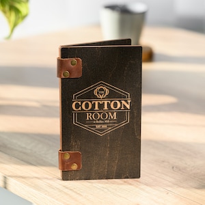 May include: A dark brown wooden menu cover with leather straps and brass accents. The cover features a gold-colored logo with the text "The Cotton Room at Belfast Mill Est 2016".