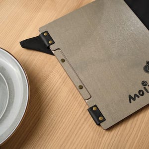 Grey wood-textured Moi cover alongside a white ceramic plate on a wooden table. Metal hinge hardware on the spine. Simple, clean composition. The white plate and grey cover create a very neutral, refined table setting. No cutlery, no props, just the cover and the plate. This minimalist styling choice suits the Mori brand aesthetic perfectly — Japanese design sensibility applied to the product photography as well as to the product itself.