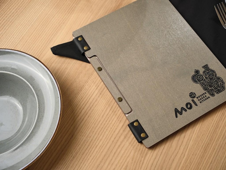 Grey wood-textured Moi cover alongside a white ceramic plate on a wooden table. Metal hinge hardware on the spine. Simple, clean composition. The white plate and grey cover create a very neutral, refined table setting. No cutlery, no props, just the cover and the plate. This minimalist styling choice suits the Mori brand aesthetic perfectly — Japanese design sensibility applied to the product photography as well as to the product itself.