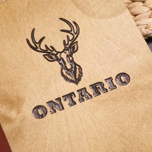Close-up of the "Ontario" stag logo engraving on the natural plywood surface. The antlers are detailed and the text sits cleanly below. The engraving depth is clearly visible — the burned lines are recessed into the plywood surface rather than just marked on top of it. The wood grain texture behind the illustration adds to the natural quality of the finished product. This is the best single shot for communicating engraving quality to a buyer considering a custom logo on this plywood finish.