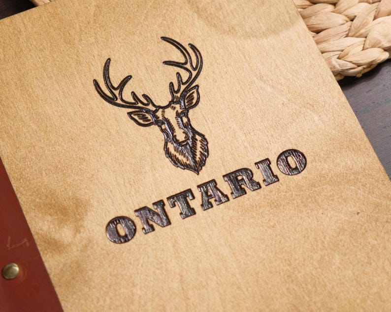 Close-up of the "Ontario" stag logo engraving on the natural plywood surface. The antlers are detailed and the text sits cleanly below. The engraving depth is clearly visible — the burned lines are recessed into the plywood surface rather than just marked on top of it. The wood grain texture behind the illustration adds to the natural quality of the finished product. This is the best single shot for communicating engraving quality to a buyer considering a custom logo on this plywood finish.