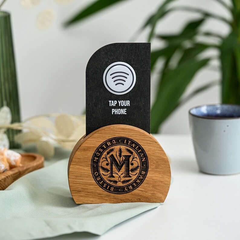 Custom NFC menu stand with black vertical display panel featuring Spotify icon, Tap Your Phone instruction and NFC symbol on a natural oak semicircle wood base with laser engraved Maestro Italian restaurant circular badge logo displayed on a white table with green plant background. The bold black panel and warm oak semicircle base create a distinctive premium branded contactless NFC digital menu accessory for Italian restaurants and upscale dining venues.