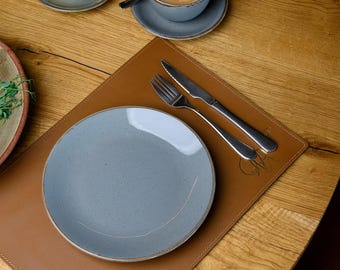 Könnte beinhalten: Ein braunes Leder-Platzset mit einem grauen Teller, einer Gabel und einem Messer. Das Platzset hat ein dezentes Prägemuster.