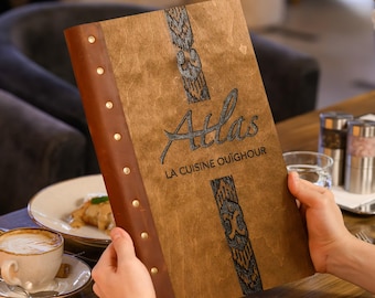 Puede incluir: Una funda de menú rectangular de madera con un lomo de cuero marrón asegurado por remaches metálicos. El frente presenta el texto grabado "Atlas" y "LA CUISINE OUÏGHOUR" con patrones verticales decorativos.