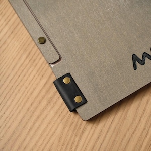 Back of the grey wood-textured Mori cover showing the script logo and a small circular seal or stamp mark below it. Metal hinge hardware on the corners. Wooden surface background. This back-cover shot is a useful addition to a product listing — it shows there's branding on the back as well as the front, and the small seal detail adds authenticity to the custom order. The engraving quality is consistent front and back, which confirms it's not just surface decoration.