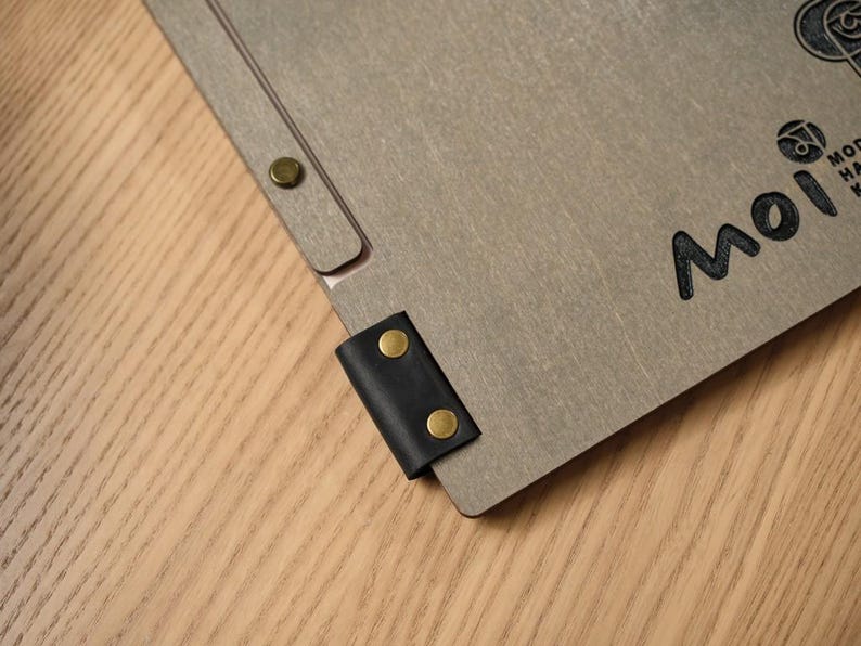 Back of the grey wood-textured Mori cover showing the script logo and a small circular seal or stamp mark below it. Metal hinge hardware on the corners. Wooden surface background. This back-cover shot is a useful addition to a product listing — it shows there's branding on the back as well as the front, and the small seal detail adds authenticity to the custom order. The engraving quality is consistent front and back, which confirms it's not just surface decoration.