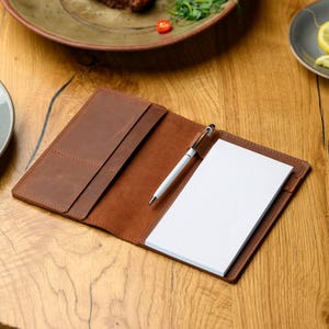 May include: A brown leather check presenter with a white notepad and a silver and white pen. The check presenter is open, revealing the notepad and pen. The leather has a textured appearance.