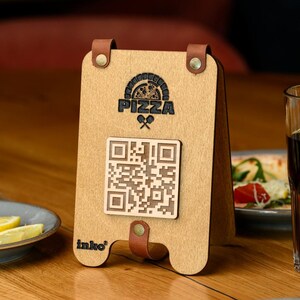 May include: Wooden pizza menu holder with a QR code and the word "PIZZA" in black. The holder has a light brown finish with brown leather accents and a logo that says "inko".