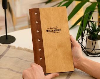 Puede incluir: Un menú de madera con lomo de cuero marrón y remaches blancos. El menú presenta el texto "The Moose & Mountie, Whitehorse, Yukon" en una fuente decorativa. El menú está abierto y sostenido por una persona.