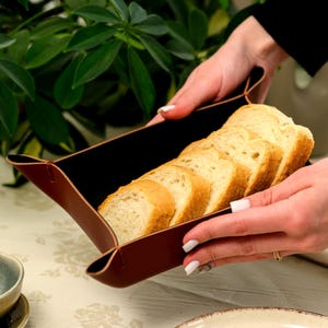 Könnte beinhalten: Ein rechteckiges Tablett aus braunem Leder mit gefalteten Ecken, das mehrere Scheiben Weißbrot enthält. Das Tablett hat eine glatte, dunkelbraune Außenseite und ein kontrastierendes Inneres und wird von zwei Händen gehalten.