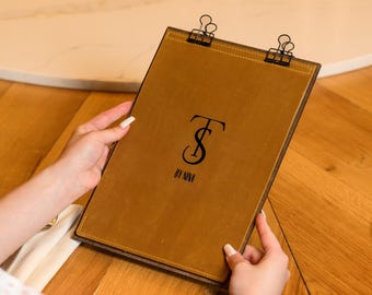 May include: A rectangular tan leather menu board featuring a black logo with the letters TS and the text BY NINA centered on the front. Two black metal binder clips are attached to the top edge, securing the leather panel to a dark backing.