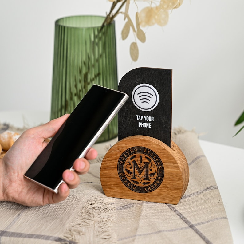 Custom NFC contactless menu stand featuring a black rectangular display panel with Spotify logo, Tap Your Phone text and NFC symbol mounted on a natural oak semicircle base with laser engraved Maestro Italian restaurant logo displayed with a hand tapping a smartphone to activate the NFC menu. The oak and black panel combination creates a premium modern contactless digital menu solution for Italian restaurants, wine bars and upscale dining venues.