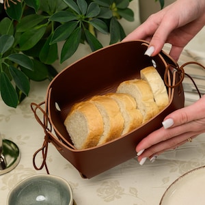 May include: A dark brown, rectangular, open-top container made of a smooth, flexible material, featuring thin cord ties at the corners. The container is filled with several slices of light-colored, crusty bread with a soft, airy interior texture.