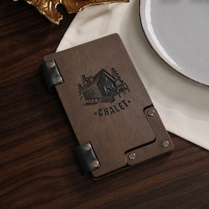 May include: A dark brown wooden menu holder with a black leather strap and metal rivets. The holder features a cabin illustration and the word "CHALET" in black. The holder is on a wooden surface.