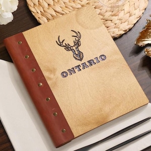 May include: Wooden menu cover with a brown leather spine and brass rivets. The cover features a deer head graphic and the word "ONTARIO" in a decorative font. The menu is resting on a white napkin with black cutlery.