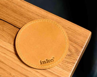 May include: A round, tan leather coaster with stitched edges rests on a wooden surface. The coaster features the brand name "inko" in a small, embossed font. The coaster is designed to protect surfaces from drink condensation.