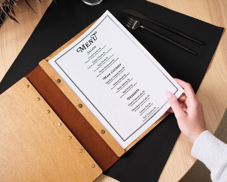 Hands flip through interior pages of the natural plywood cover on a table. The leather spine is on the right edge.  This action shot shows the plywood cover handling repeated page-turning without any issues. The pages stay well-aligned inside the cover and the leather spine shows no sign of stress from being repeatedly opened and closed. For a product that gets used dozens of times per service, this kind of durability demonstration is more convincing than any written description could be.