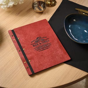 Puede incluir: Un menú de madera roja con el texto "North Branch Bar & Grill Drink Menu" se muestra sobre una mesa de madera clara. Una servilleta negra, un cuenco azul y cubiertos dorados también están sobre la mesa. El menú está encuadernado con una tira negra y remaches de latón.