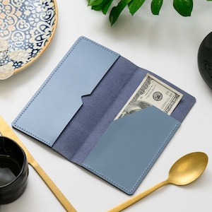 May include: Open light blue leather wallet with a dark blue interior. A one hundred dollar bill is visible in the front pocket. The wallet is open, revealing the interior pockets and stitching.
