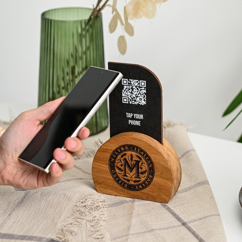 Hand holding a smartphone tapping the NFC display panel on a custom oak and black Maestro Italian restaurant menu stand on a white table with plant background demonstrating the NFC contactless activation of digital menu access. The practical NFC tapping demonstration highlights how this stand enables instant digital menu access for guests through smartphone NFC technology in Italian restaurants and upscale dining venues.