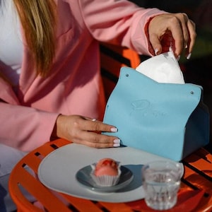 May include: A pale blue tissue box with the word "Plume" embossed on the front. The tissue box is open, revealing white tissues. A small plate with a pink pastry and a glass of water are on a table.