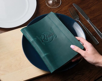 May include: A teal leather menu cover with the word "Romeo" embossed in a circle. The menu is on a dark blue plate and a wooden table.