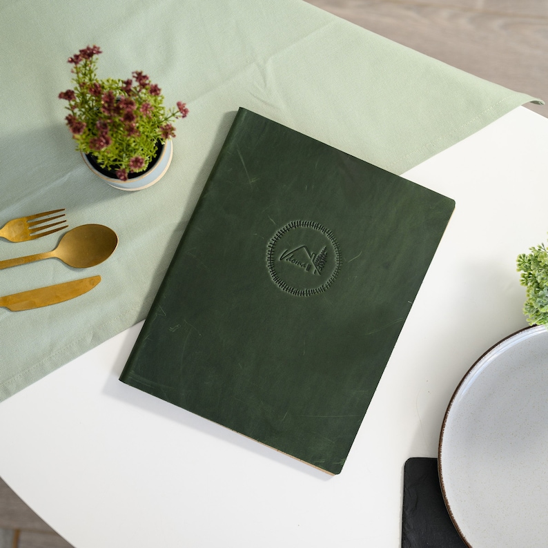 Dark forest green Crazy Horse leather menu cover with custom embossed logo placed flat on a modern white restaurant table next to gold cutlery, a decorative ceramic dish and green plant. Angled lifestyle product photo highlighting the genuine aged leather texture, rich dark green color and snap button closure strap. Designed for cafes, bistros and upscale fine dining restaurants requiring branded menus.