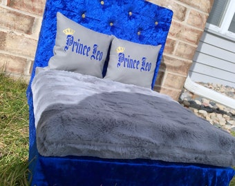 Könnte beinhalten: Ein blaues Samt-Hundebett mit einer grauen Kunstfellmatratze und zwei grauen Kissen mit goldenen Kronen und dem in Blau gestickten Text "Prince Leo".
