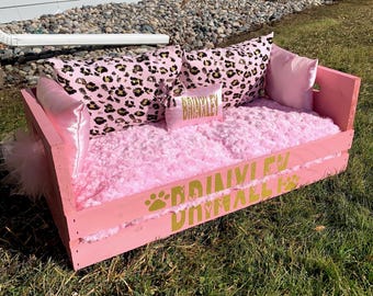 May include: A pink wooden pet bed with a fluffy pink interior and several pillows. The pillows include a leopard print design and a small pillow with the name "BRAINLEY" in gold lettering. The bed is decorated with gold paw prints and text.