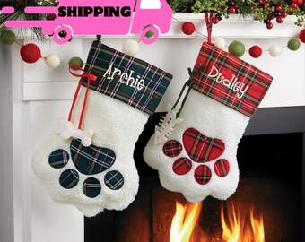 May include: Two personalized Christmas stockings with a paw print design. The stockings are red and green plaid with white fur and have the names "Archie" and "Dudley" embroidered on them.