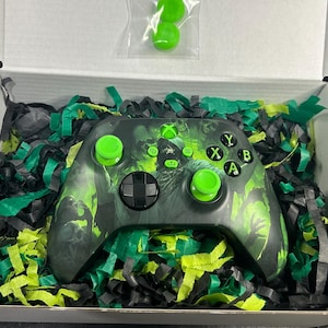 May include: A black and neon green video game controller featuring a graphic print of a hooded figure, nestled in a white box with black and green shredded paper filler. Two additional neon green thumbstick covers are included in a small bag.
