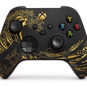 May include: A black wireless video game controller featuring a gold-coloured graphic of a large ocean wave and a mountain landscape. The controller includes a central Xbox button, two black joysticks, a directional pad, and four coloured action buttons.