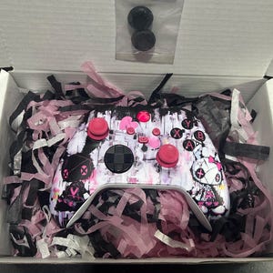 May include: A custom video game controller featuring a white, black, and pink graphic print with a character illustration, pink thumbsticks, and standard buttons, nestled in a white box with pink and black shredded paper filler and two spare parts.