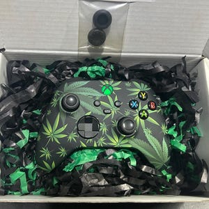 May include: A black video game controller featuring a green leaf pattern, standard buttons, and a glowing green Xbox logo. The controller is nestled in black and green shredded paper inside a white box, with two small black thumbstick covers included.
