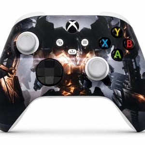 May include: A wireless video game controller featuring a dark, high-contrast graphic print of a robotic figure with glowing orange accents. The device includes two light grey analogue sticks, a black directional pad, and four coloured action buttons.