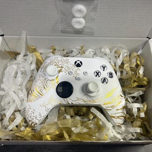 May include: A white wireless game controller featuring a gold wave pattern across the front shell. The controller includes standard buttons labelled A, B, X, and Y, a black directional pad, and two white thumbsticks. Two white thumbstick covers are included.