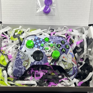 May include: A custom video game controller featuring a graphic print of a comic book character with green hair and a purple suit, accented with bright green thumbsticks and buttons, packaged in a white box with shredded paper and two purple caps.