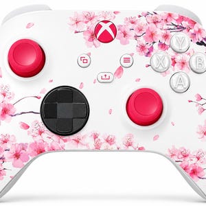 May include: A white wireless video game controller featuring a pink cherry blossom floral pattern across the front. The device includes two bright pink thumbsticks, a black directional pad, white action buttons labelled A, B, X, Y, and a central Xbox logo button.