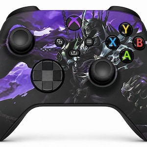 May include: A black video game controller featuring a purple and black graphic of a knight in armour with chains. The controller includes a central Xbox button, two black joysticks, a directional pad, and four coloured action buttons labelled A, B, X, and Y.
