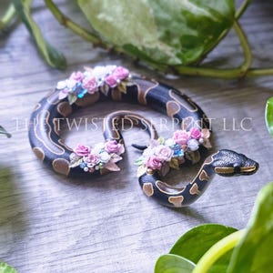 May include: A decorative snake figurine with a black and brown pattern, adorned with pink and white floral accents and blue gems. The snake is coiled and has a detailed head. The text "THE TWISTED SERPENT LLC" is visible.