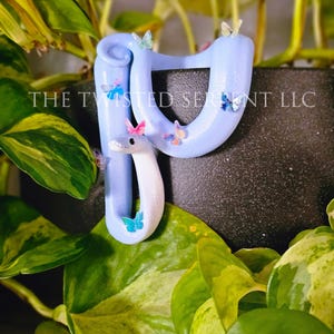 May include: A light blue snake-shaped sculpture with a white head and colorful butterfly accents. The sculpture is decorated with small, multi-colored butterflies and the text "THE TWISTED SERPENT LLC". The sculpture is set against a backdrop of green foliage.