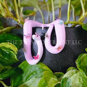 May include: A light pink sculpture of a snake with a white head and small, colorful butterflies. The snake is decorated with butterflies in shades of pink, orange, and purple. The text "THE TWISTED SERPENT LLC" is visible. The sculpture is surrounded by green foliage.