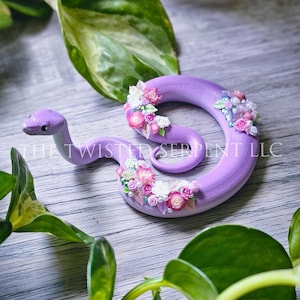 May include: A lavender-colored snake figurine adorned with clusters of pink, white, and pearl-like flowers. The snake is coiled, with its head raised. The text "THE TWISTED SERPENT LLC" is visible. Green leaves surround the figurine.