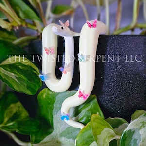 May include: A cream-colored snake sculpture adorned with colorful butterfly accents. The snake is positioned in a decorative arrangement with green foliage and the text "THE TWISTED SERPENT LLC".