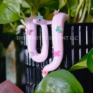 May include: A light pink snake figurine adorned with small, colorful butterflies. The snake is positioned among green foliage and a black container. The text "THE TWISTED SERPENT LLC" is visible on the side.