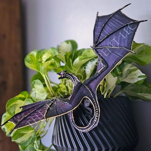 May include: A detailed, dark purple dragon figurine with gold accents and outstretched wings. The dragon is perched above a black, geometric planter filled with green and yellow pothos plants. The dragon's wings are a deep purple with gold trim.