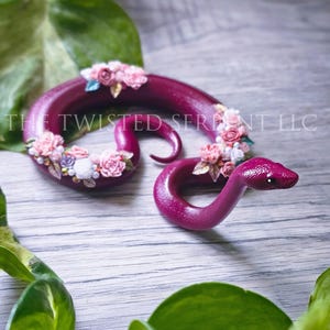 May include: A handcrafted, deep magenta snake figurine adorned with delicate pink and white floral accents. The snake is coiled and features detailed scales and a realistic head. The text "THE TWISTED SERPENT LLC" is visible.